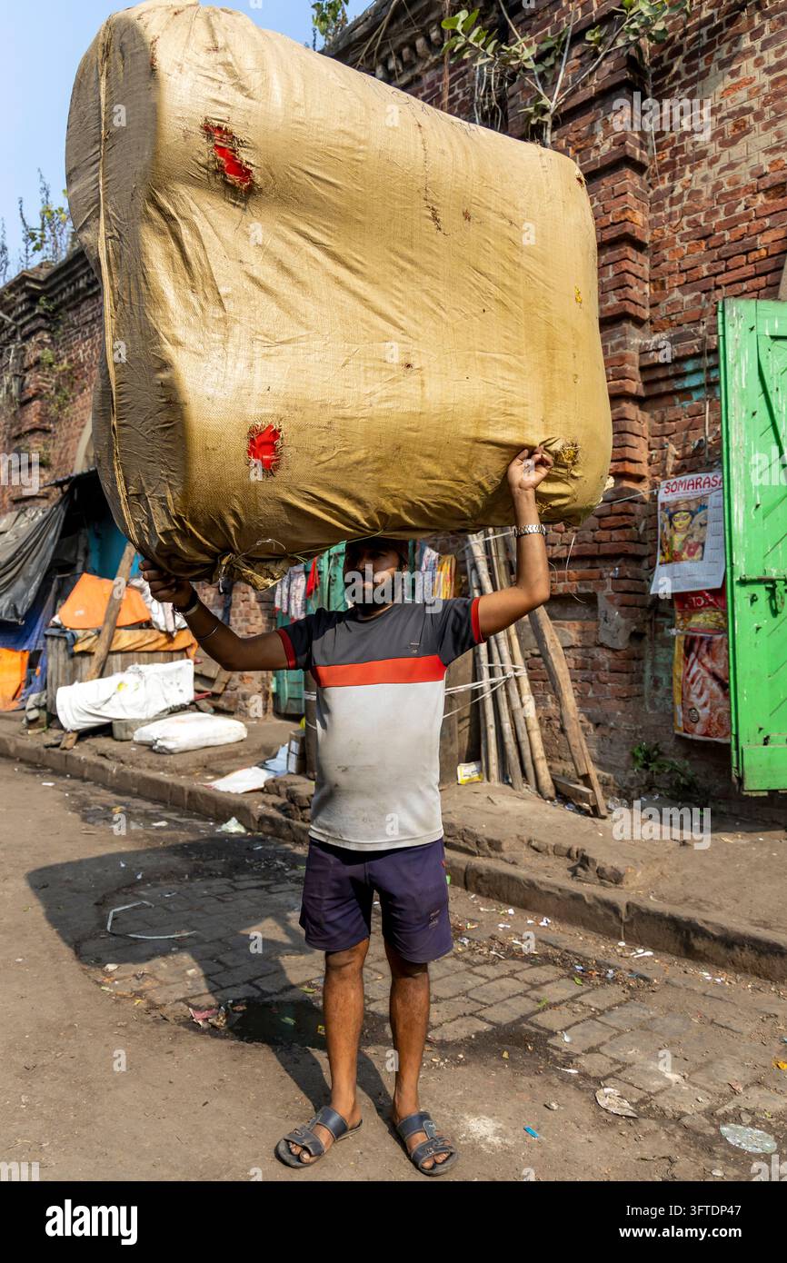 Porter in una camicia a righe e pantaloncini si trova in un viale tranquillo bilanciando un pacco sovradimensionato avvolto in tessuto marrone, Bengala Occidentale, Howrah, India Foto Stock