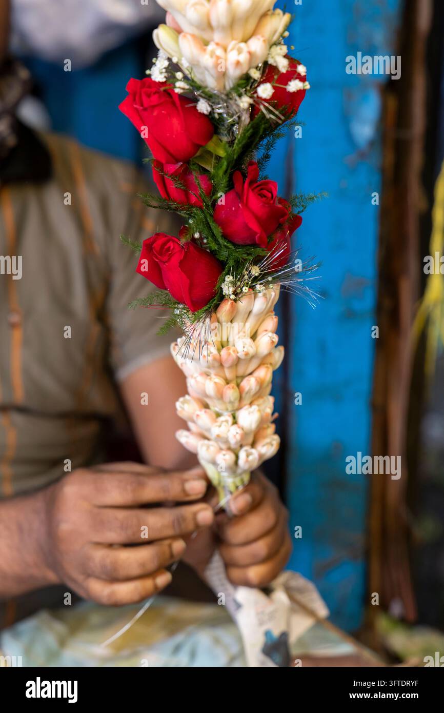 Prima vista di mani che lavorano su una lunga ghirlanda di rose rosse e foglie verdi al mercato dei fiori, Bengala Occidentale, Kolkata, India Foto Stock