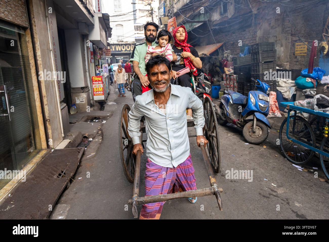 Un uomo sorridente con una camicia bianca tira una rickshaw che trasporta un uomo, una donna con una sciarpa rossa e un bambino lungo una stradina, Bengala Occidentale, Kolkata, India Foto Stock
