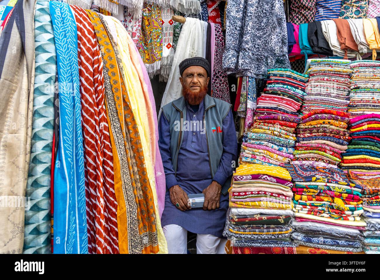 Un uomo barbuto con un cappello a testa di morto e un gilet è seduto al suo banco tra tessuti stampati appesi e pile di stoffa piegata, Bengala Occidentale, Kolkata, India Foto Stock