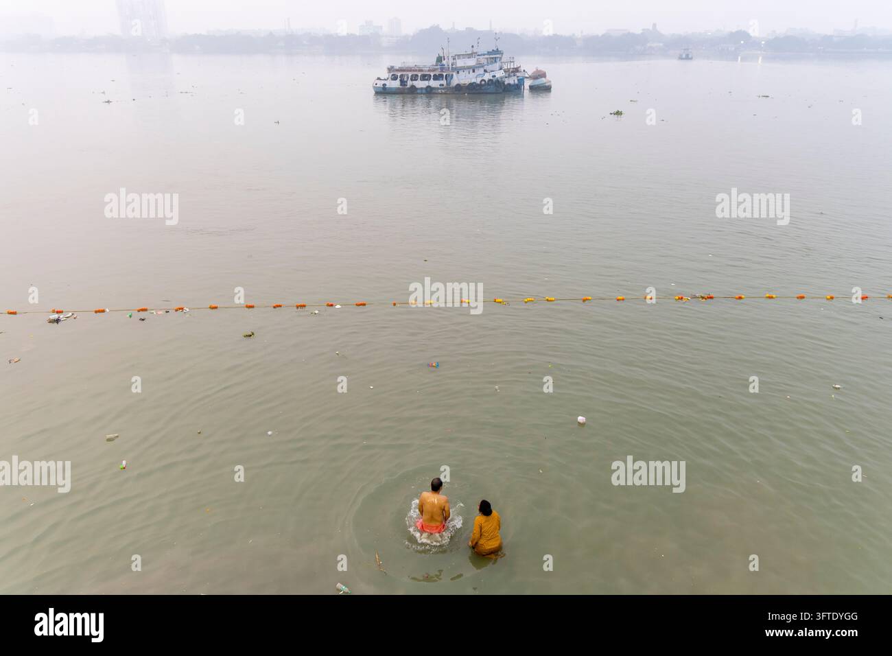 Due persone si bagnano nell'acqua bassa di un ampio fiume nebbioso mentre una barca si trova lontano, Bengala Occidentale, Kolkata, India Foto Stock