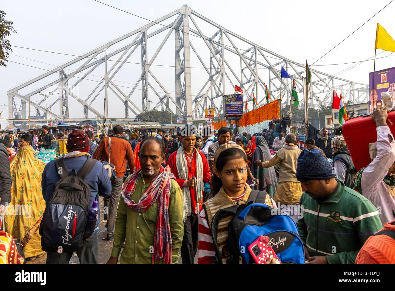 Una folla indaffarata in abiti invernali che porta zaini cammina lungo una strada con un grande ponte a tralicci d'acciaio sullo sfondo, Bengala Occidentale, Kolkata, India Foto Stock