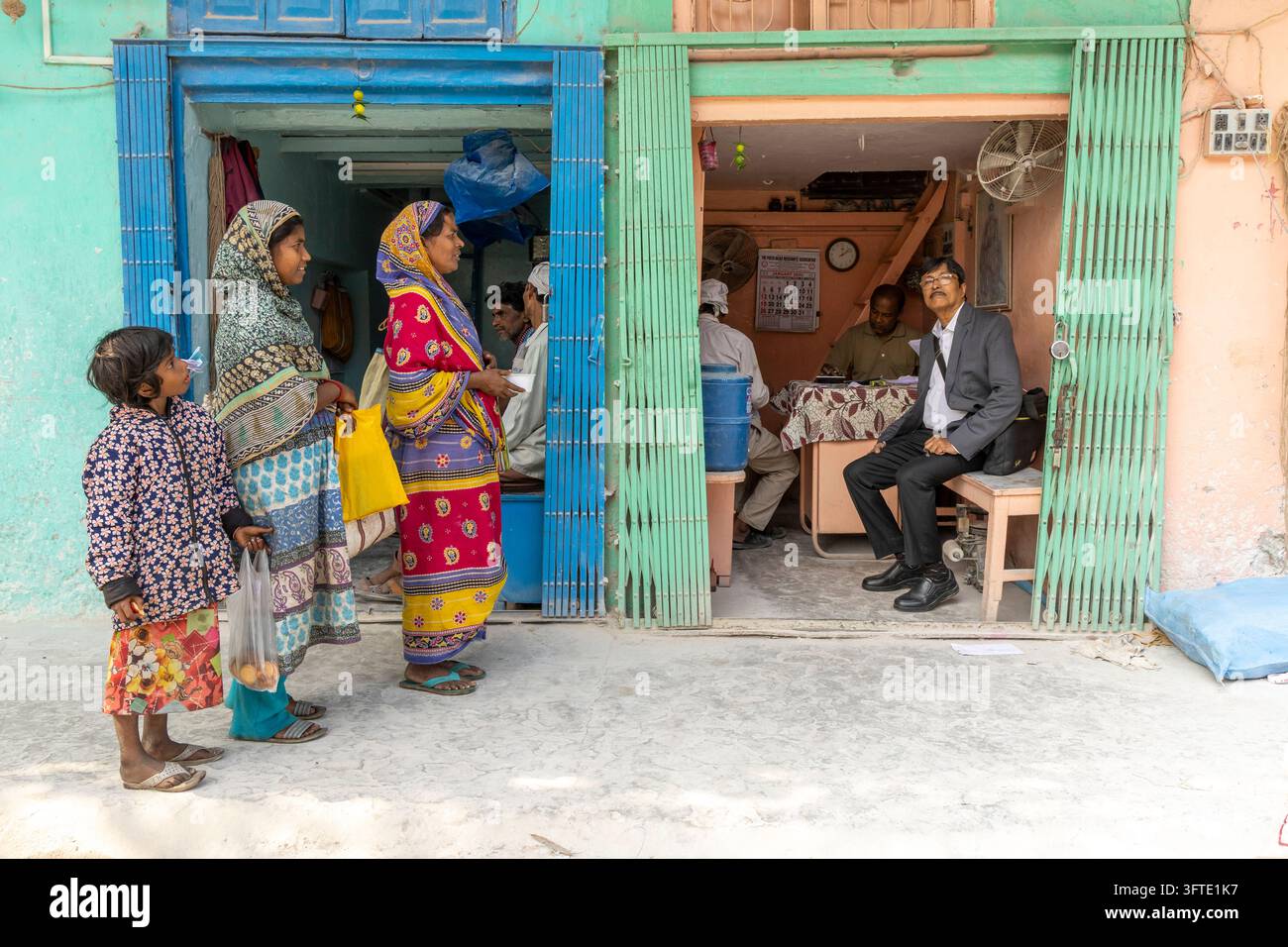 Due donne in sari colorati e due ragazze aspettano fuori da un edificio dipinto di blu e verde mentre uomini seduti osservano, Bengala Occidentale, Kolkata, India Foto Stock