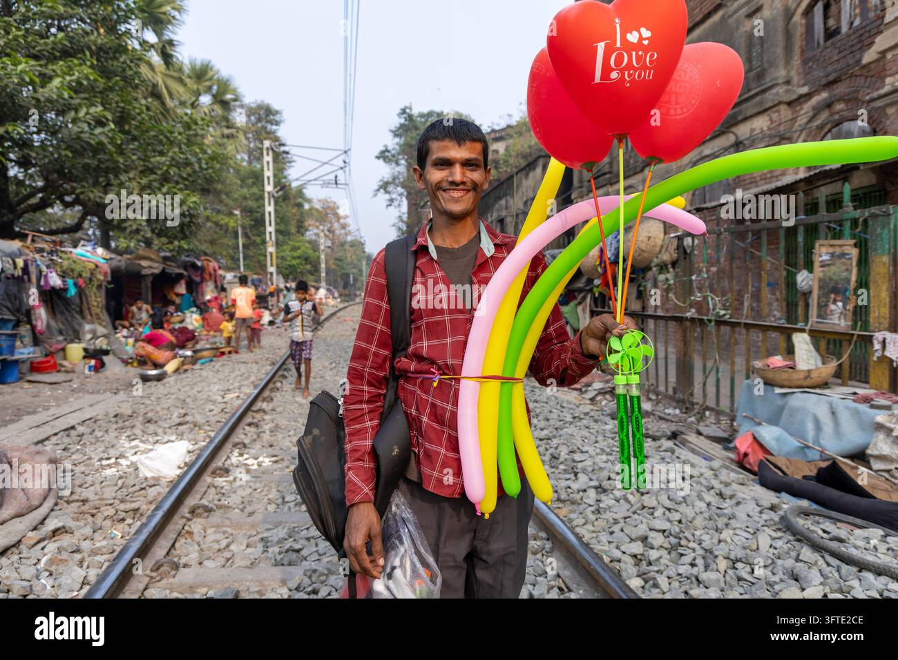 Venditore allegro con uno zaino e palloncini a forma di cuore rosso si trova in fila vicino a famiglie sedute vicino ai loro rifugi, Bengala Occidentale, Kolkata, India Foto Stock