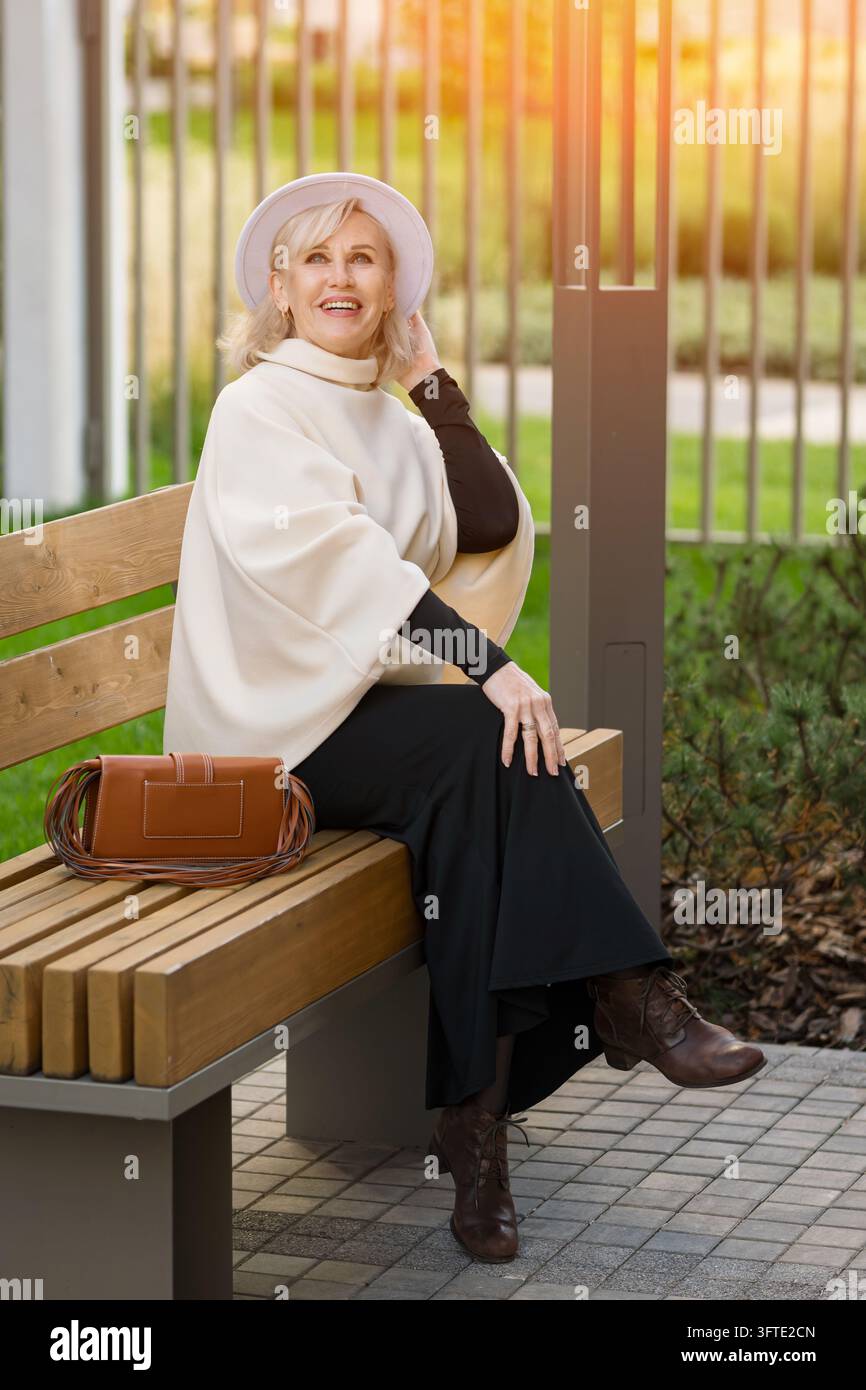 Donna matura felice seduta comodamente su panchina di legno con borsa in pelle in un giardino luminoso. Donna elegante tiene il suo cappello mentre si gode il tempo libero all'aperto Foto Stock