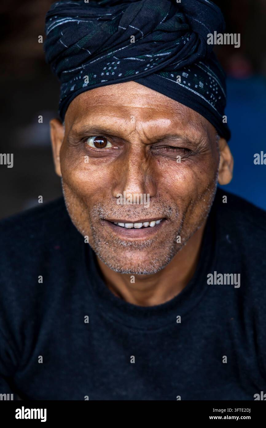 Un uomo con barba grigia e turbante scuro annodato sorride e fa l'occhiolino alla telecamera in un ritratto verticale stretto, Bengala Occidentale, Kolkata, India Foto Stock