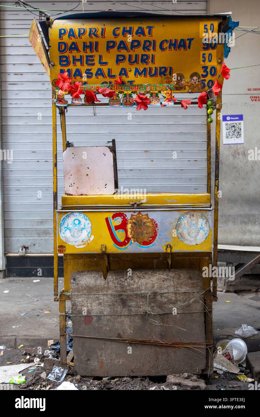 Bancarella di cibo in legno giallo vuota con una lista dei prezzi dipinta a mano per snack e fiori di ibisco su un marciapiede sporco, Bengala Occidentale, Kolkata, India Foto Stock
