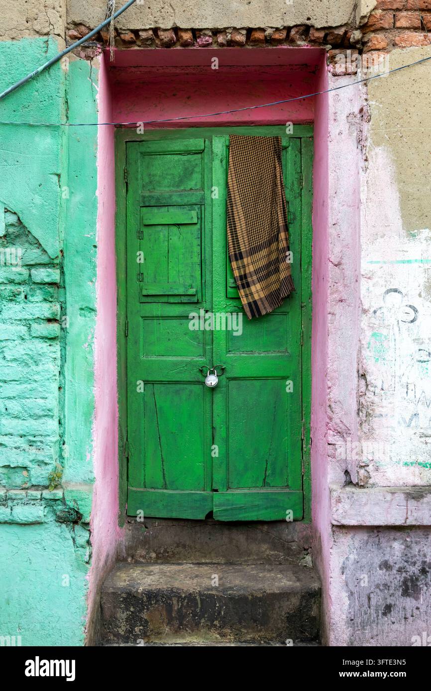 Una porta di legno verde serrata con un lucchetto in una nicchia dipinta di rosa in un muro verde menta con un panno a quadri appeso sopra, Bengala Occidentale, Kolkata, India Foto Stock