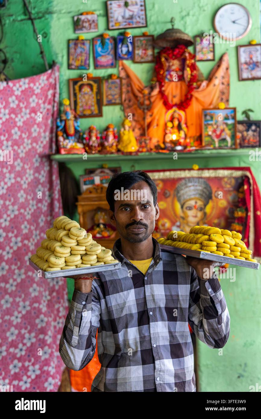 Venditore di dolci si trova in un angolo del negozio presentando due vassoi di metallo pieni di dolci gialli rotondi sotto immagini di divinità, Bengala Occidentale, Kolkata, India Foto Stock