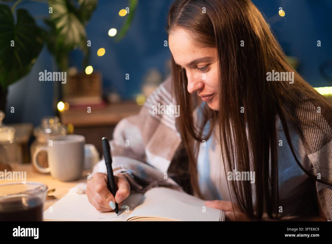 Journaling e pianificazione, giovane donna avvolta in una calda coperta a quadri che scrive nel diario a casa. Concetto di journaling, pianificazione, stile di vita accogliente e organizzazione personale Foto Stock