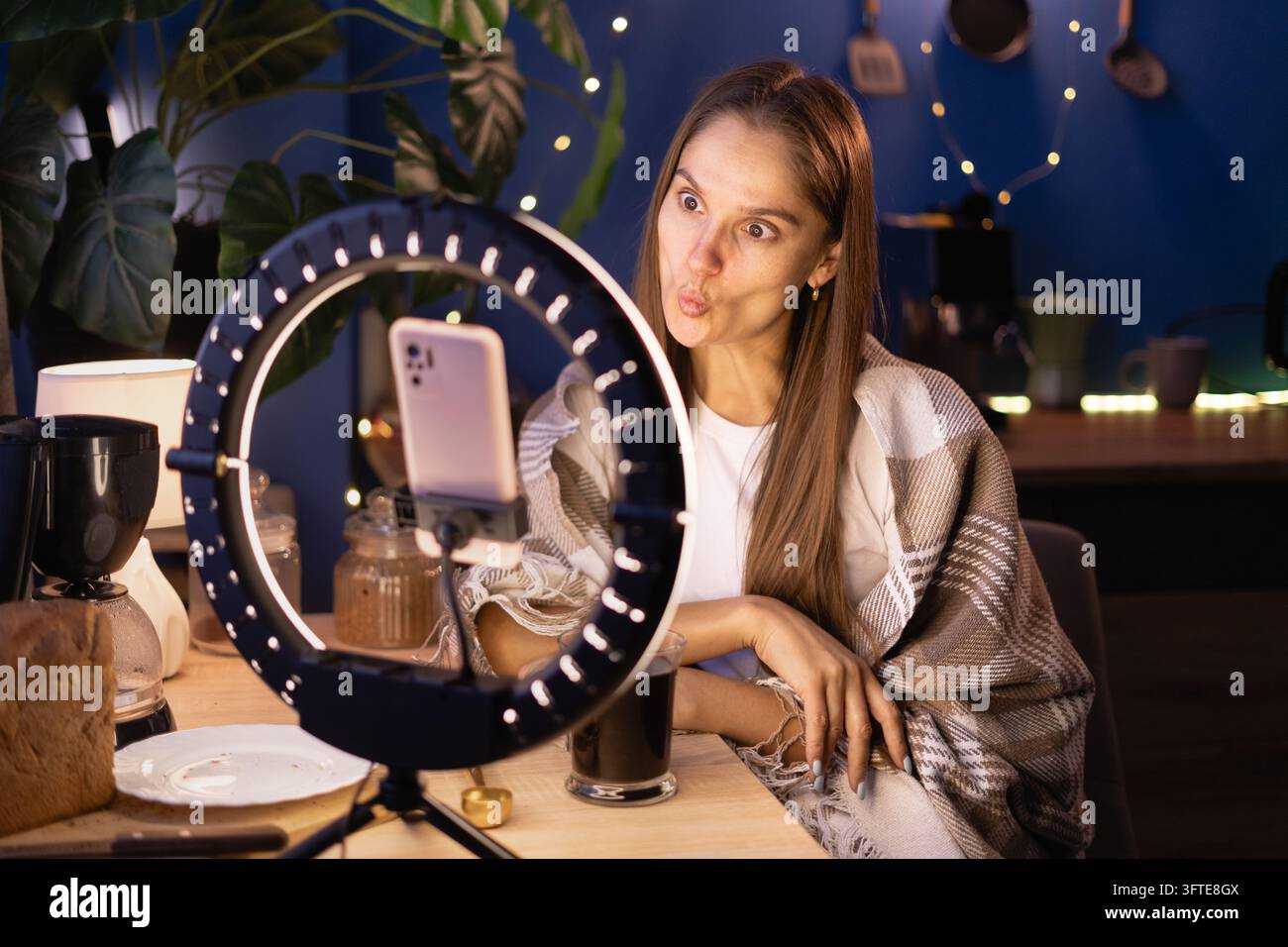 Influencer Goofy Woman che copre la produzione di coperte calde, facendo labbra a faccia di pesce, filmando contenuti video per i social media usando il telefono con anello luminoso a casa, live stre Foto Stock