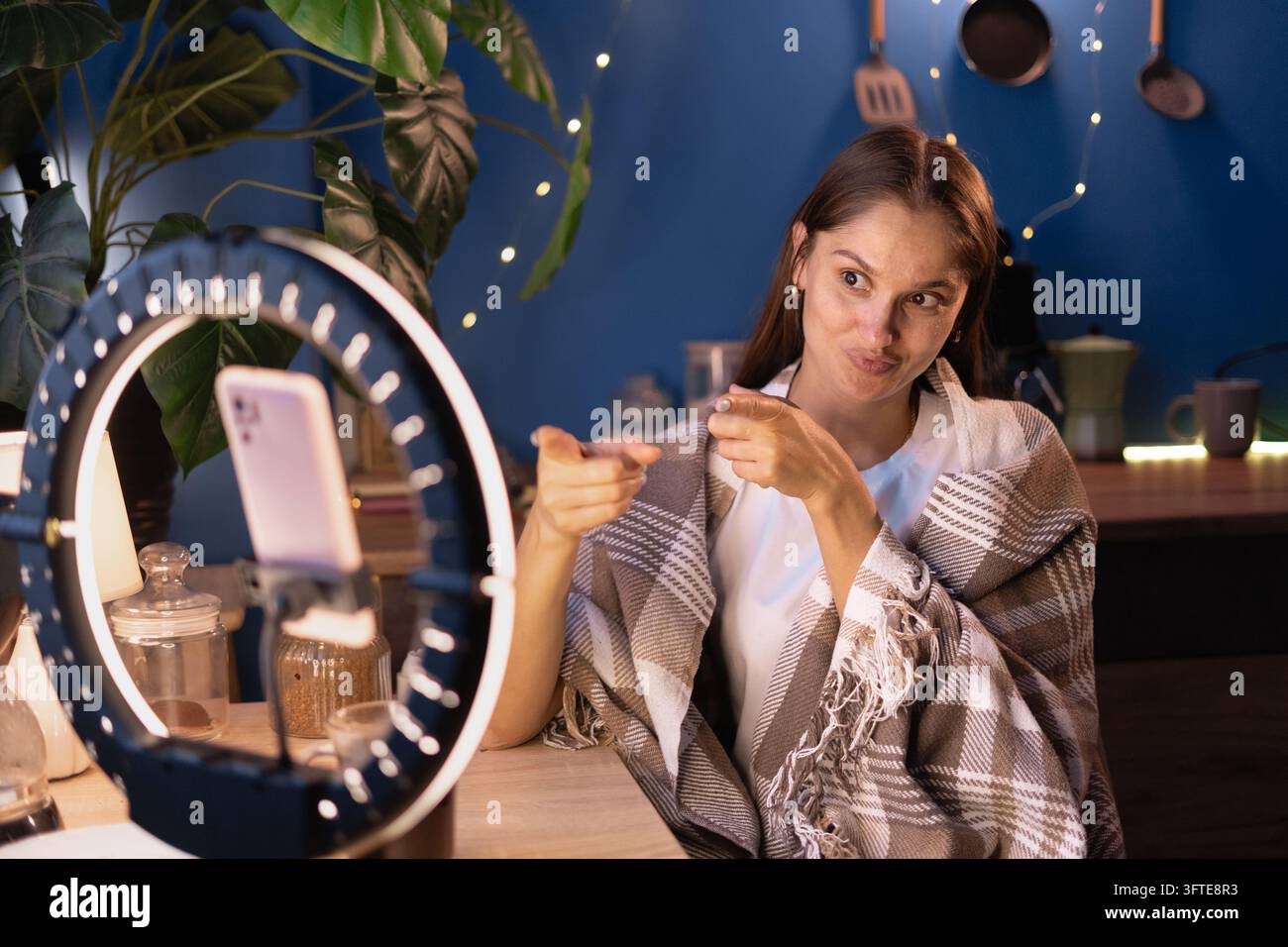 Influencer donna che copre la creazione di coperte calde, fa un gesto con le dita a forma di pistola per salutare un amico, filma contenuti video per i social media usando il telefono con anello luminoso a h Foto Stock