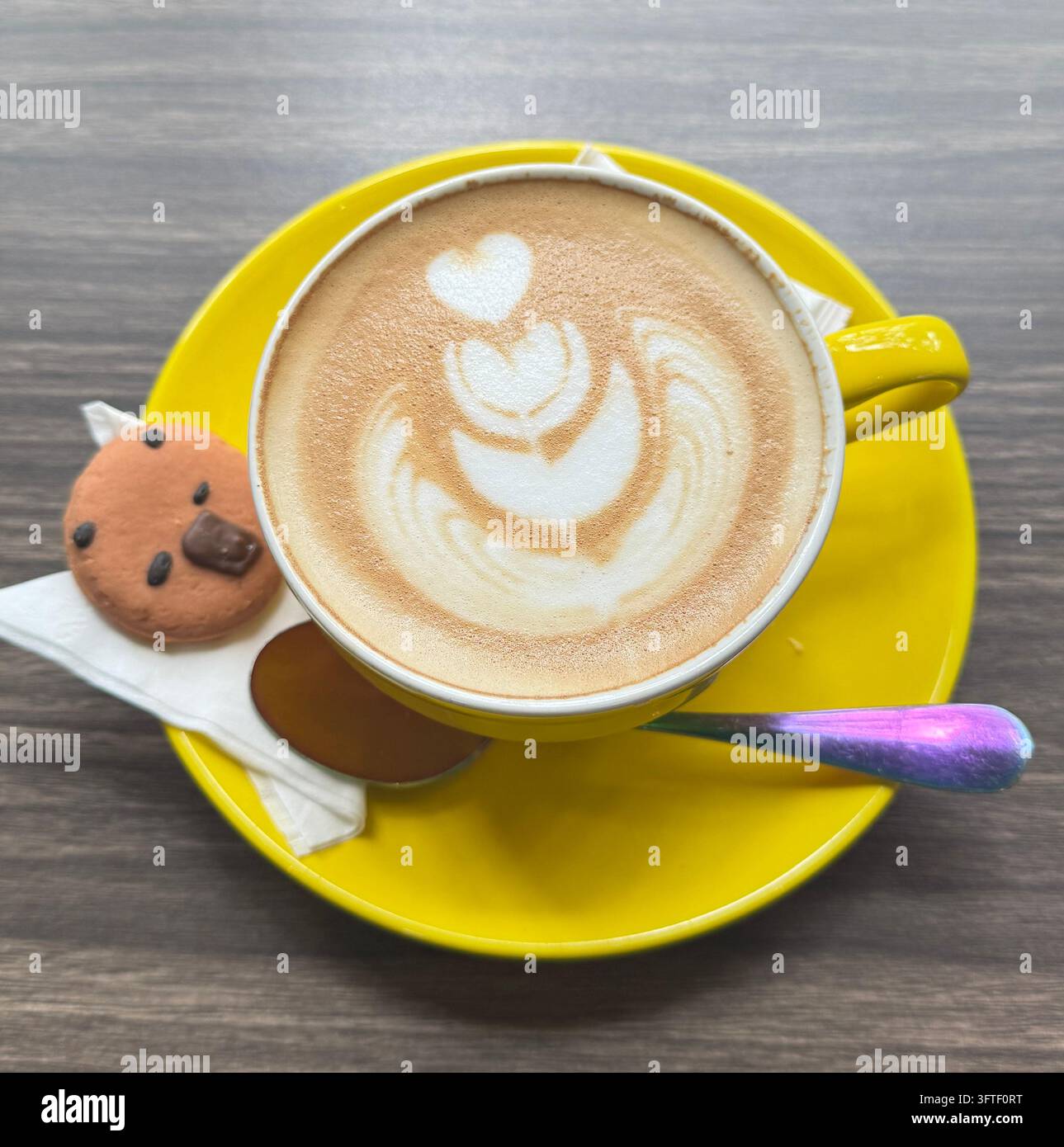 Flat lay di caffè latte art in tazza gialla con piattino e biscotto a forma di orsetto carino su tavolo di legno, vista dall'alto - Immagine stock catturata con smartphone