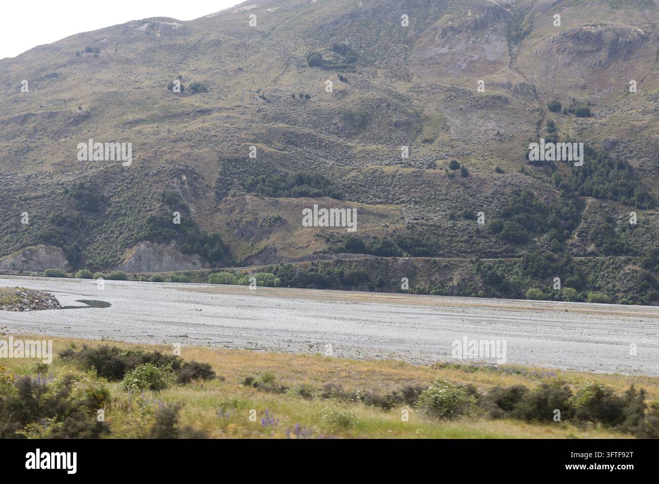 ampia valle con letto fluviale ghiaioso di fronte a grandi montagne asciutte e inclinate (grandangolo, panorama, cielo nuvoloso, vista da treno in movimento) Foto Stock