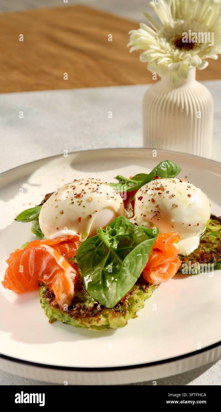 Colazione sana su un piatto piano bianco: frittelle di zucchine, uovo in camicia, salmone e foglie di spinaci. Luce naturale del mattino Foto Stock