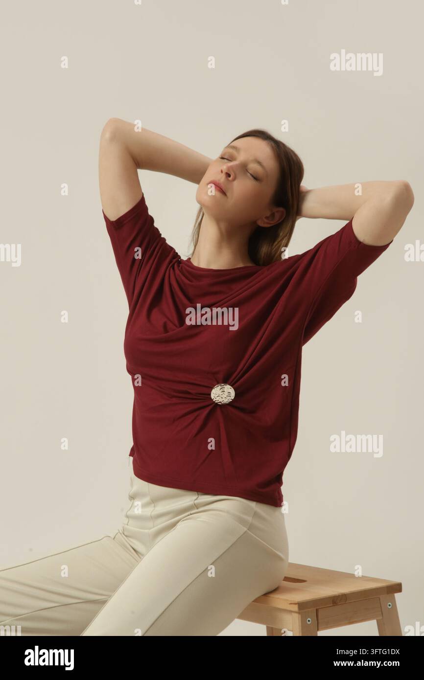 Giovane modella femminile che indossa una camicetta di cotone bordeaux con maniche a pipistrello e decorazioni metalliche, con pantaloni di cotone color crema, scatto in studio Foto Stock