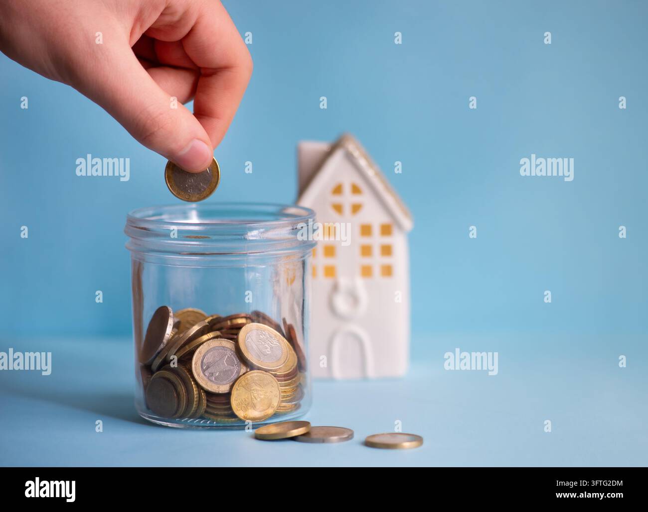 Mano che mette una moneta in euro in un barattolo di risparmio di vetro con una casa in miniatura su sfondo blu. Concetto di risparmio domestico e budget familiare. Foto Stock