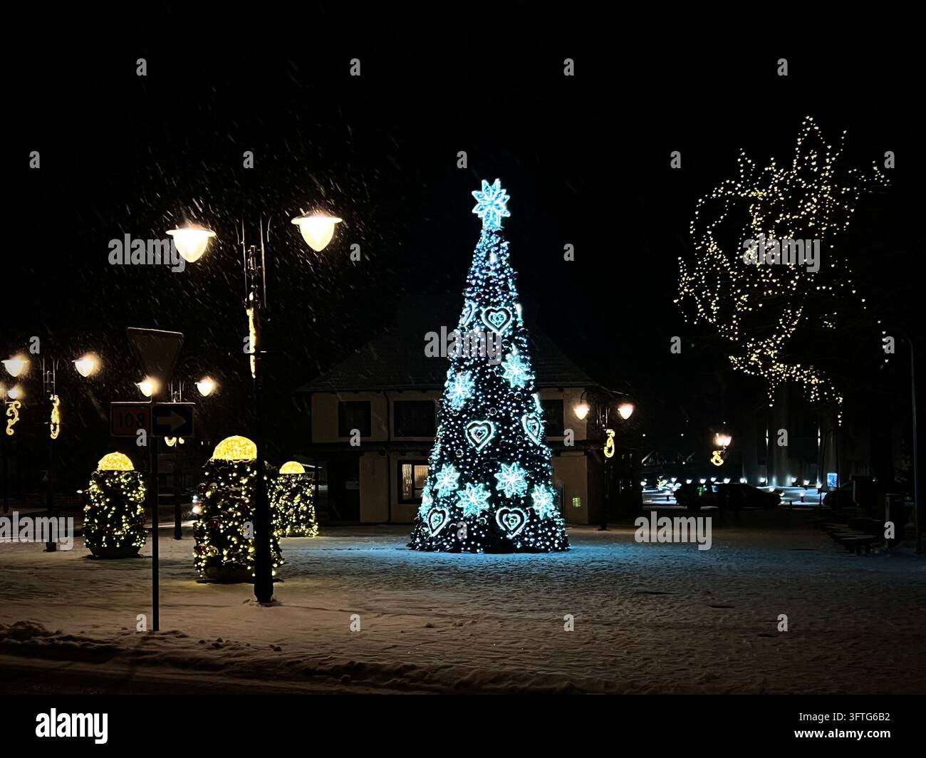 un alto albero di Natale luminosamente illuminato adornato con motivi a forma di cuore e fiocco di neve in una piazza coperta di neve a Nemenčinė durante la nevicata. Foto Stock