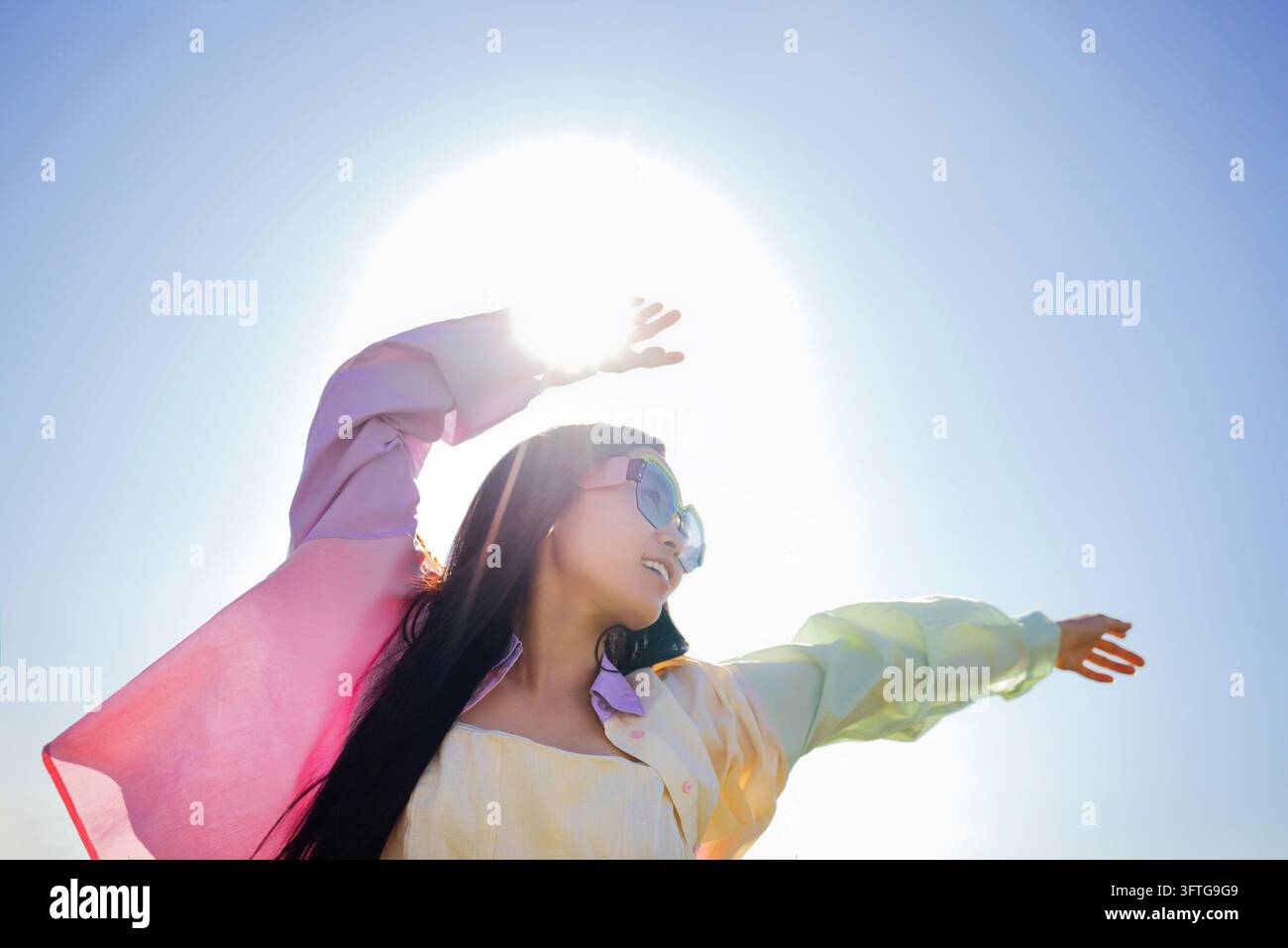 Donna con occhiali da sole alza le braccia sotto un sole luminoso, mentre maniche colorate e fluenti e lunghi capelli scuri si stagliano contro il cielo blu Foto Stock