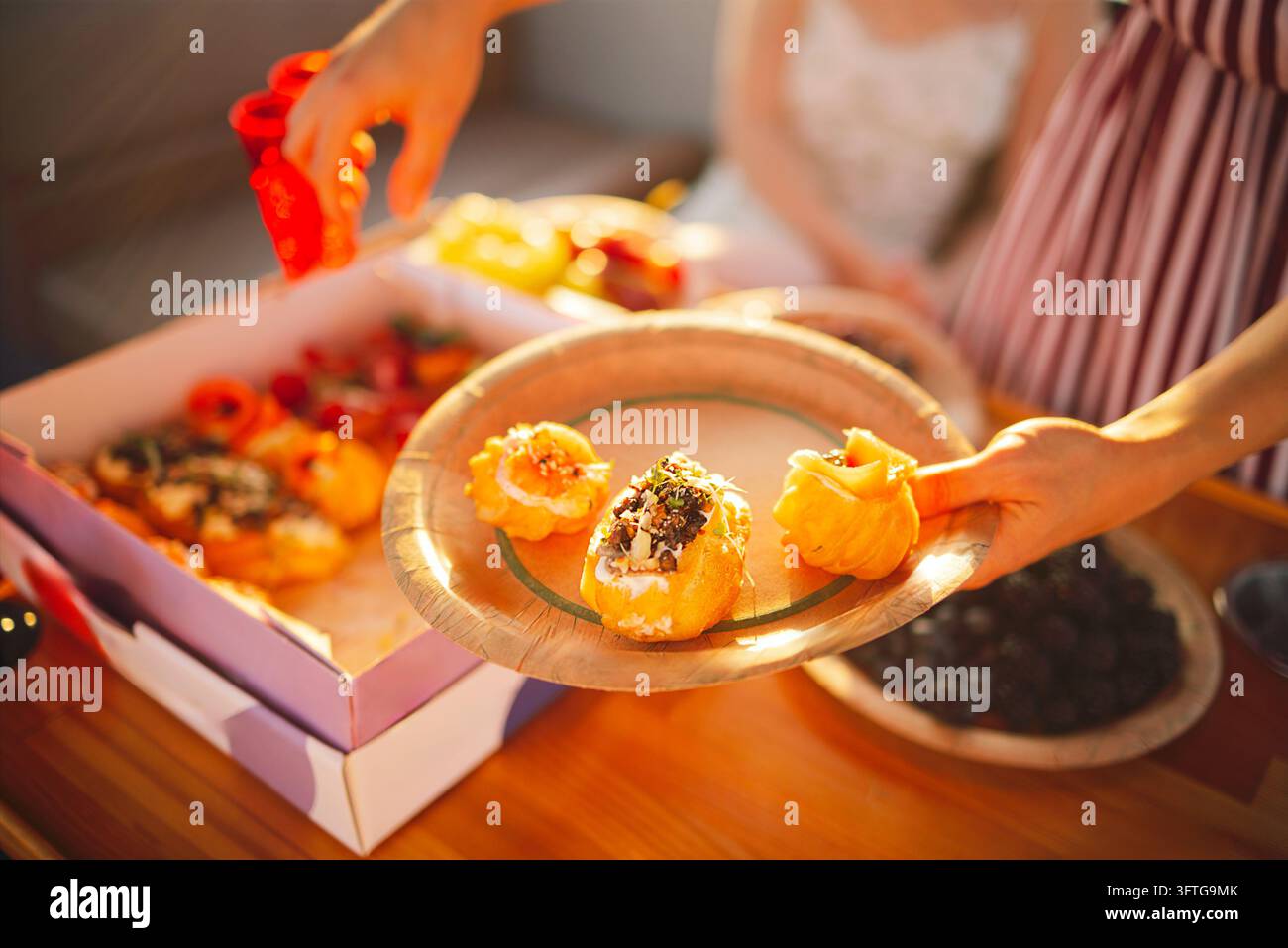 Mani dispongono vari pezzi di sushi su un piatto di vetro accanto a una scatola da asporto aperta, con ulteriori cibi e ciotole visibili su un tavolo. Golden Hour Food M Foto Stock