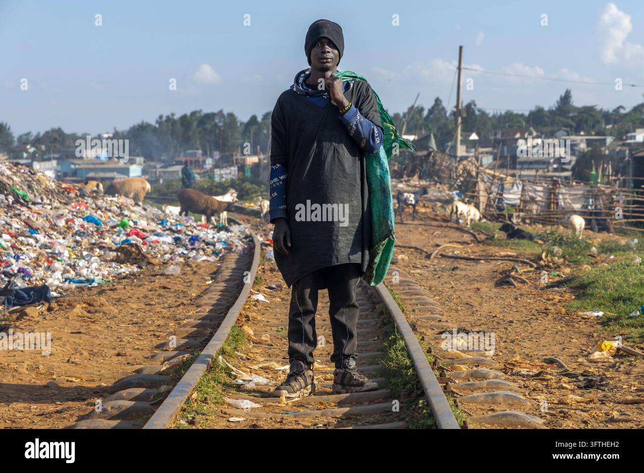 Giovane uomo con un berretto nero su binari ferroviari con un sacco verde accanto a un cumulo di rifiuti e capre a Kibera, Contea di Nairobi, Nairobi, Kenya Foto Stock