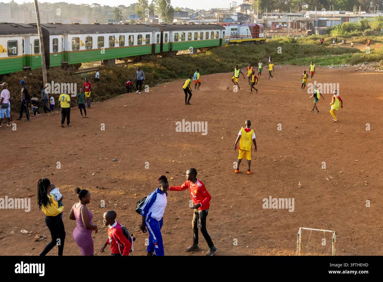 Ragazzi che giocano a calcio su un campo di terra mentre un treno pendolare verde passa dietro nell'area di Kibera, Contea di Nairobi, Nairobi, Kenya Foto Stock