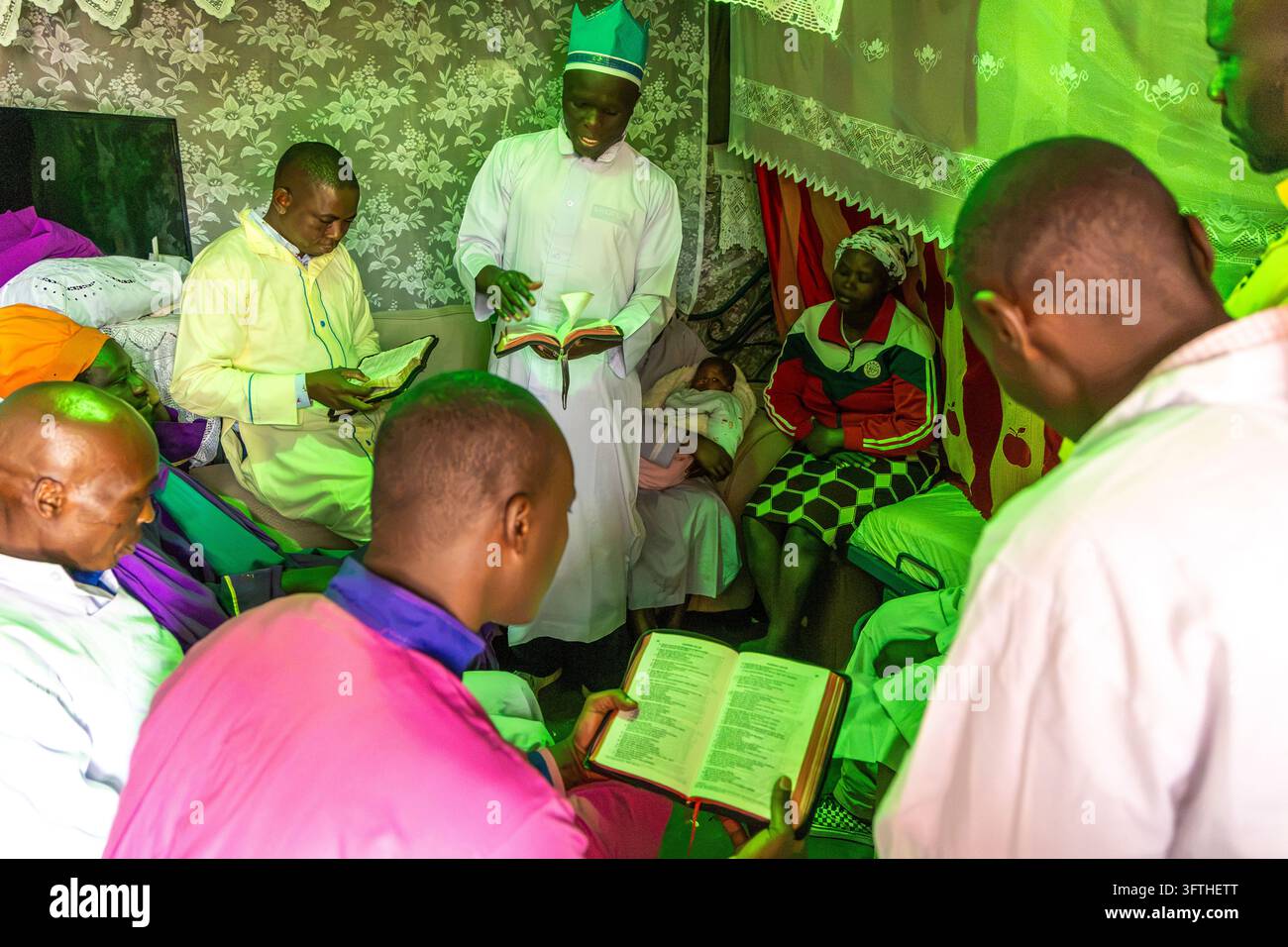 Pastore in veste bianca e cappello a corona verde che legge una Bibbia tra uomini seduti durante un battesimo a Kibera, Contea di Nairobi, Nairobi, Kenya Foto Stock