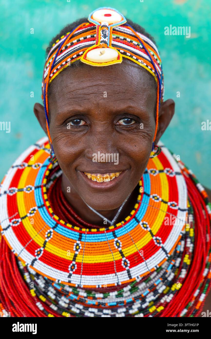 Donna sorridente della tribù Rendille con una fascia per capelli con perle e un grande colletto di perle rosse davanti a un muro verde, Contea di Marsabit, Loiyangalani, Kenya Foto Stock