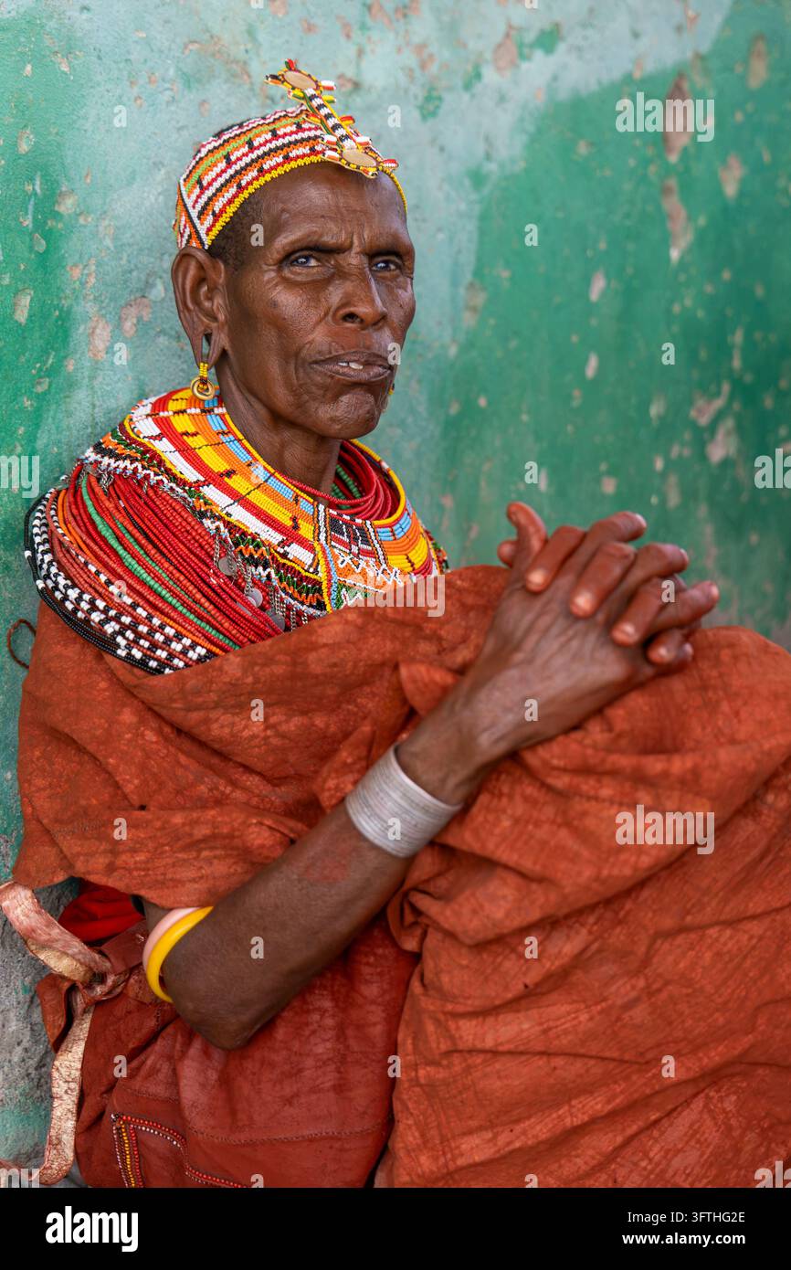 Anziana donna della tribù Rendille seduta contro un muro verde con un mantello arancione e collane di perle, Contea di Marsabit, Loiyangalani, Kenya Foto Stock