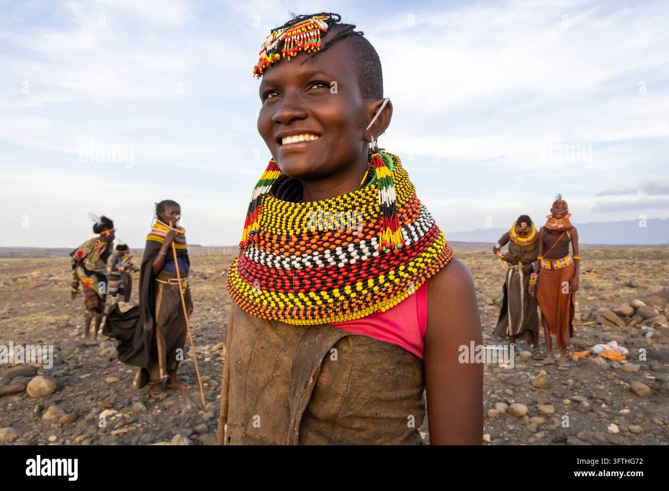 Donna sorridente della tribù Turkana con un copricapo decorato con perline e un collare a strati colorato con perline su una pianura rocciosa, Contea di Marsabit, Loiyangalani, Kenya Foto Stock