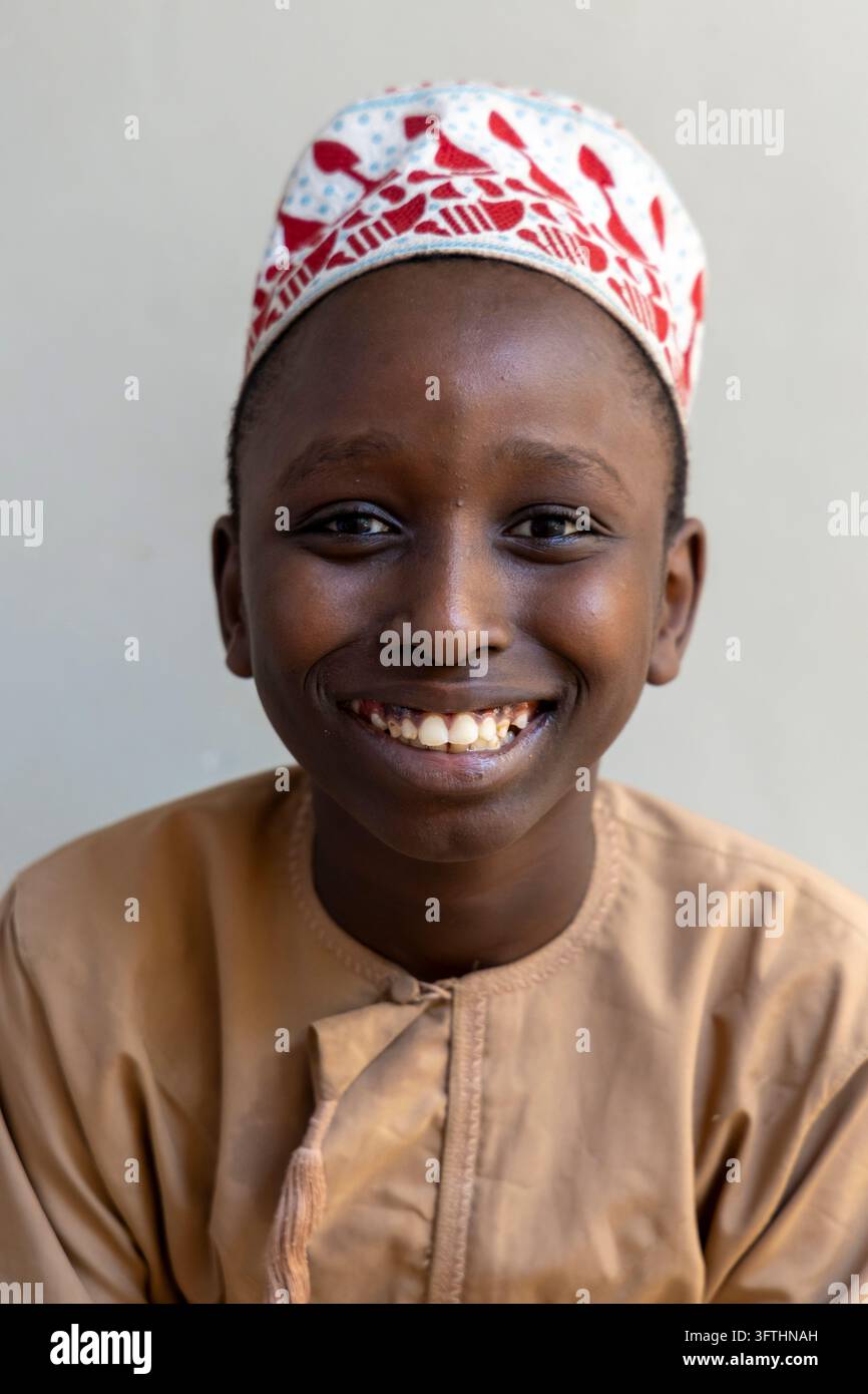 Ritratto verticale di un ragazzo che ride con una kofia ricamata rosso e bianca e una veste marrone chiaro con nappina davanti a un muro chiaro, Stone Town, Tanzania Foto Stock