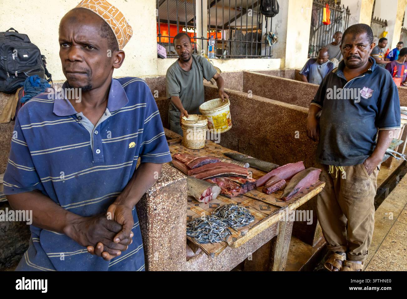 Bancarella del mercato del pesce con tre uomini accanto a un bancone piastrellato che espone tranci di tonno e piccoli pesci argentati nella sala affollata di Stone Town, Tanzania Foto Stock