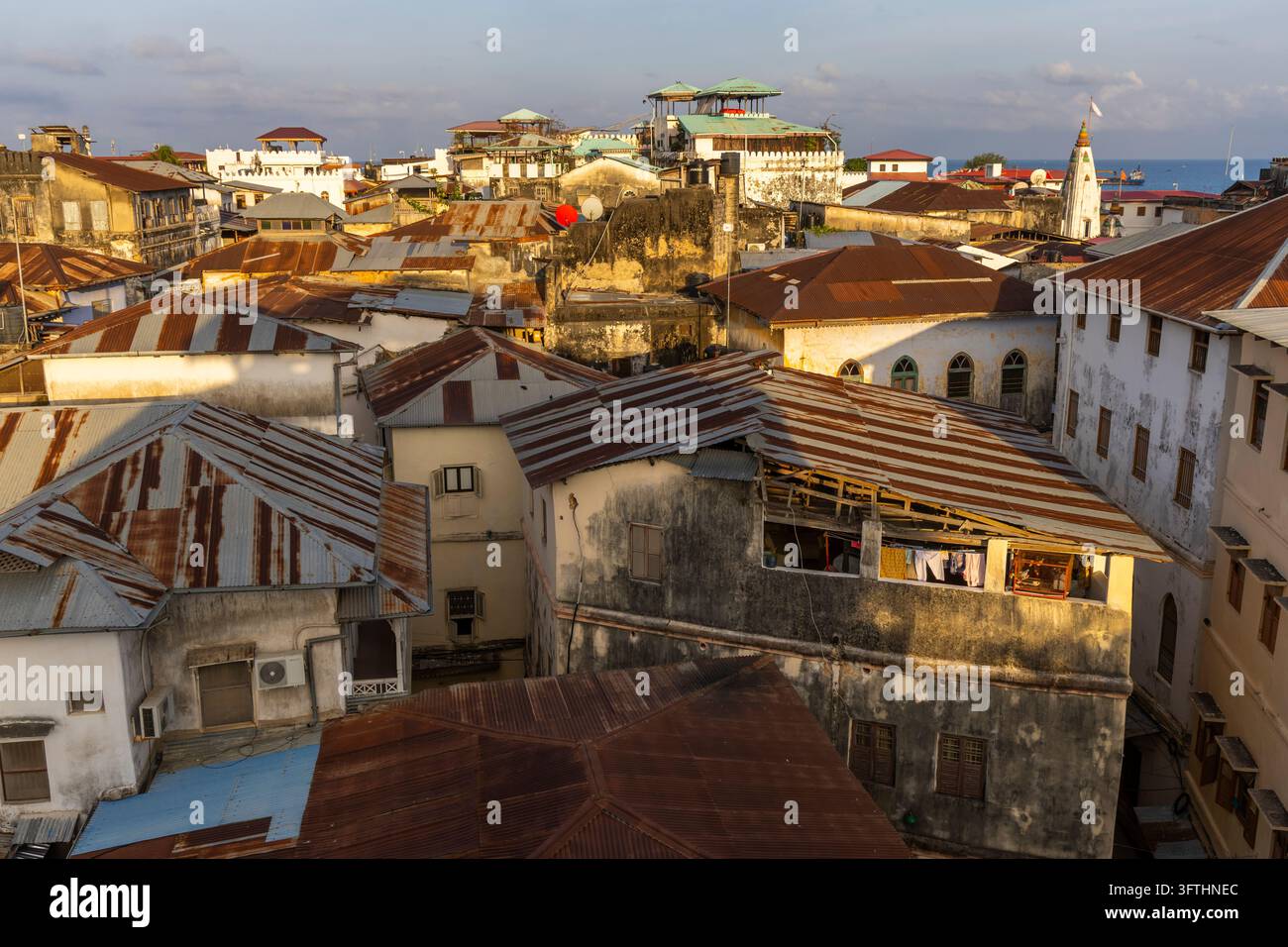 Vista ad angolo alto di tetti di ferro arrugginiti e vecchi edifici nella calda luce serale con un campanile bianco del tempio e l'oceano, Stone Town, Tanzania Foto Stock