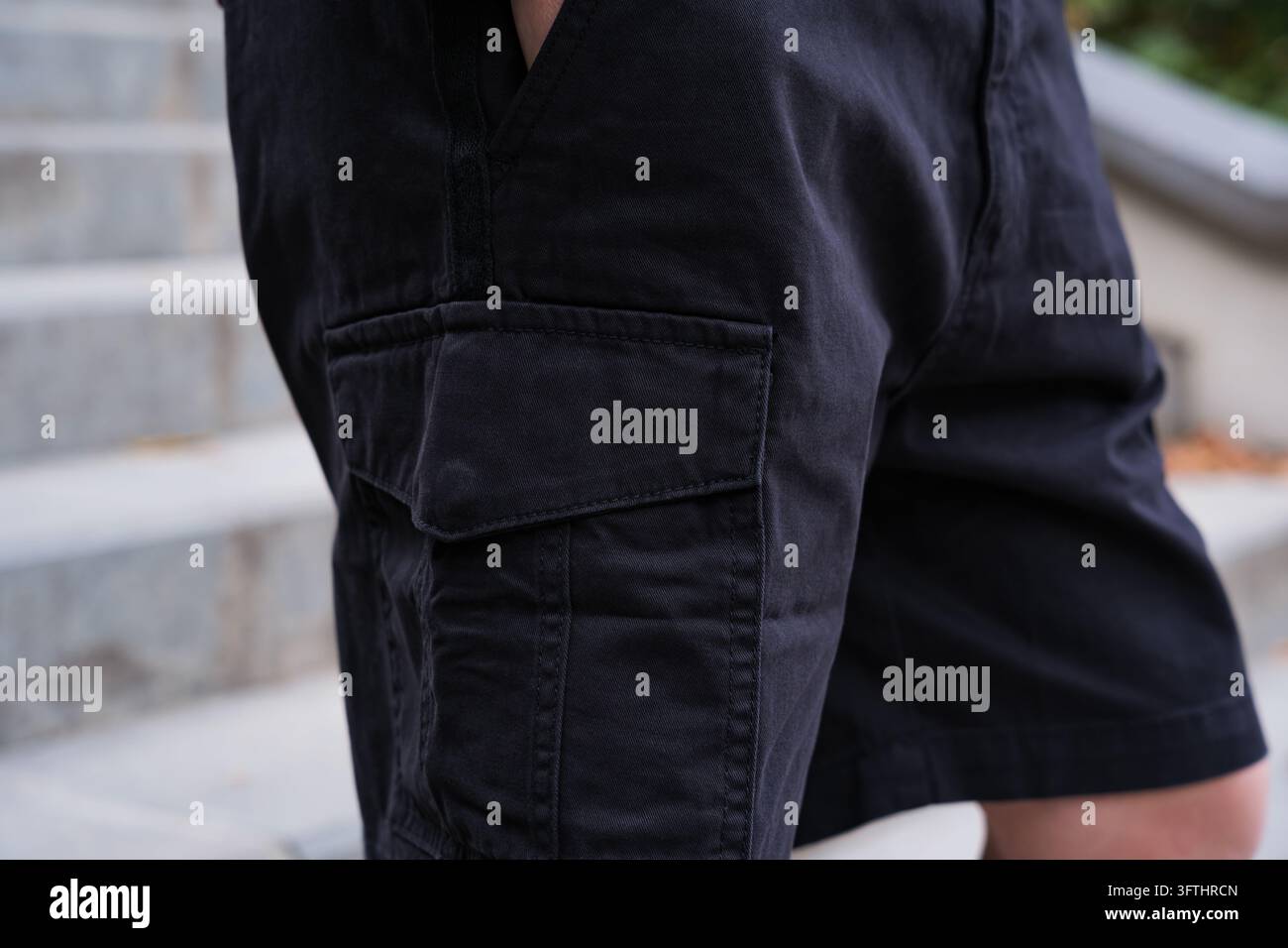 Un primo piano laterale di una giovane persona che indossa pantaloncini cargo neri con una tasca laterale e risvolto prominenti, in piedi all'aperto su gradini di cemento durante da Foto Stock
