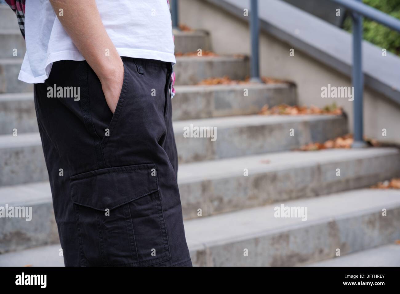 Un primo piano di lato di una giovane persona che indossa una maglietta bianca e pantaloncini cargo neri con le mani in tasca, in piedi su gradini di cemento all'aperto con f Foto Stock