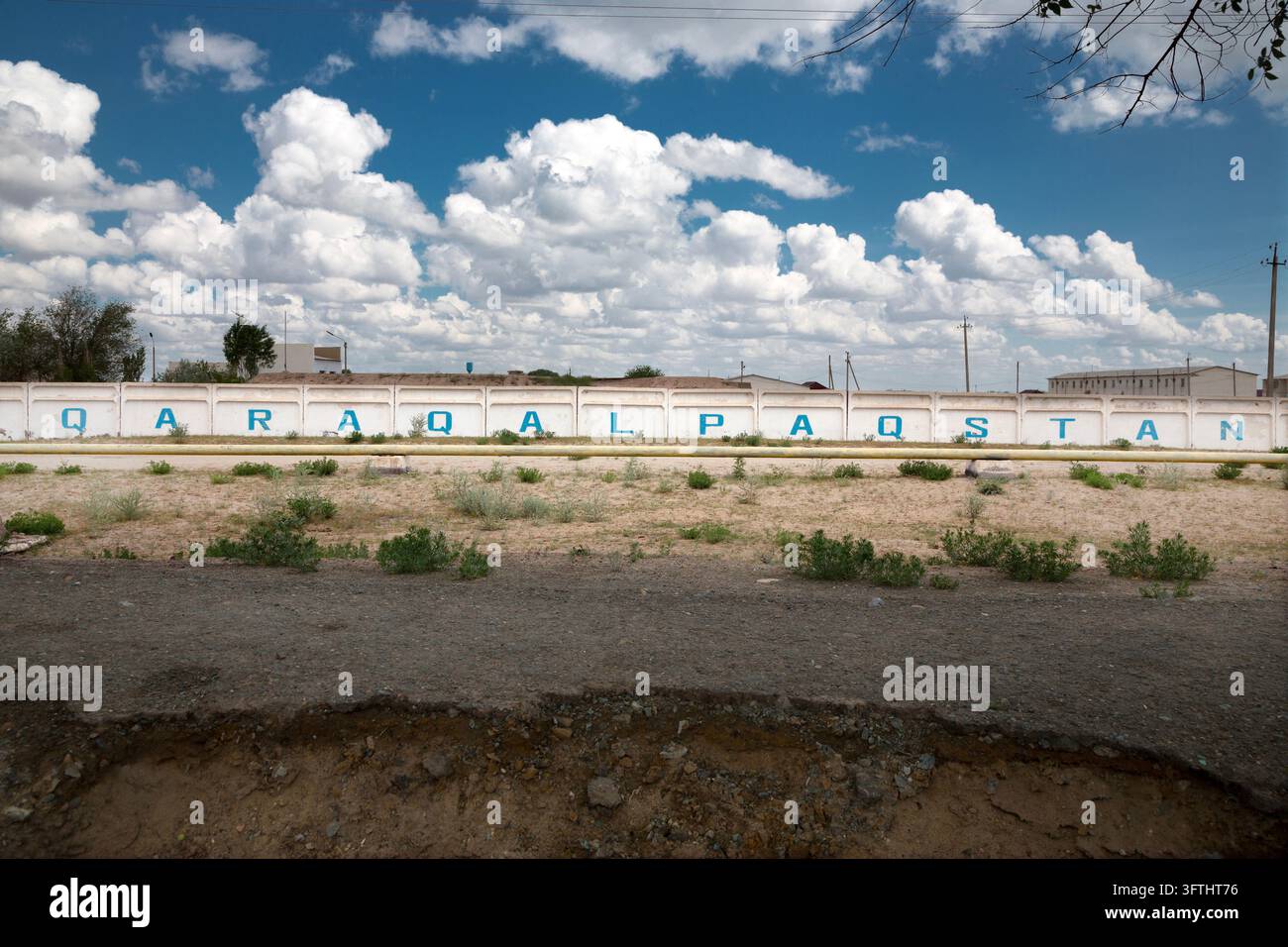 Grandi lettere blu dipinte su un lungo muro di cemento bianco accanto a un tubo del gas e a una strada sterrata sotto un cielo pieno di nuvole bianche, Moynaq, Uzbekistan Foto Stock