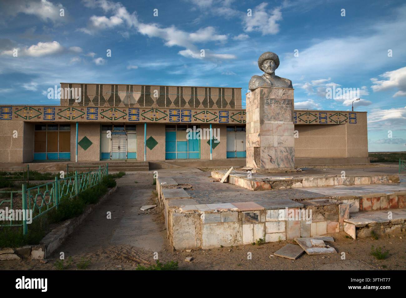 Busto in pietra di un uomo barbuto con cappello di pelliccia su un piedistallo di marmo davanti a un edificio decorato con motivi di piastrelle al crepuscolo, Moynaq, Uzbekistan Foto Stock