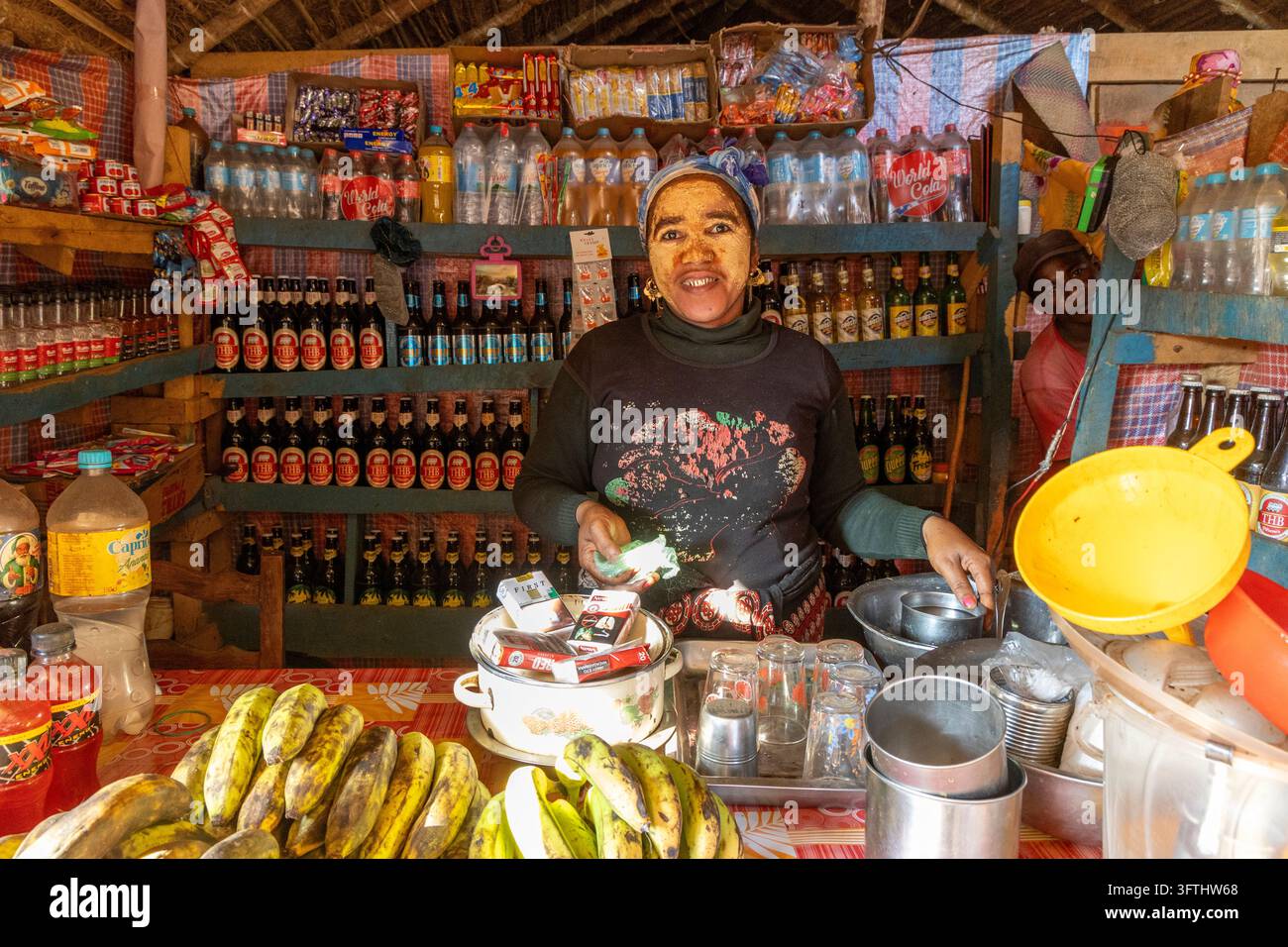 Donna che indossa una maschera facciale tradizionale gialla dietro il bancone di un piccolo negozio rifornito di bevande e banane, Menabe, Tsiribihina, Madagascar Foto Stock