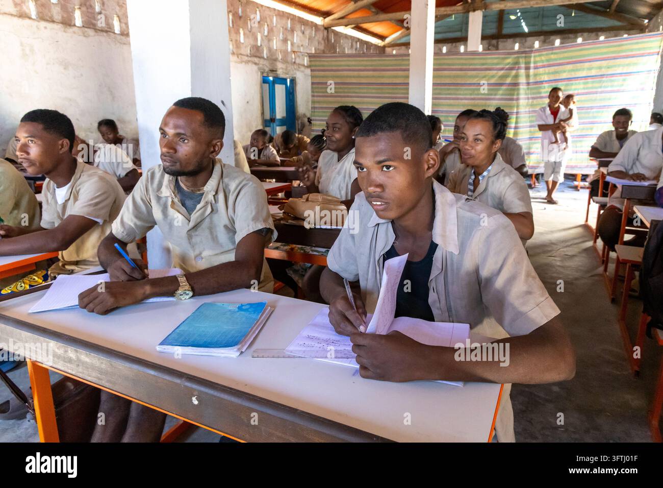 Studenti Vezo che scrivono ai banchi in un'aula all'aperto con pareti in tessuto a righe, mentre una donna tiene un bambino, Menabe, Belo sur Mer, Madagascar Foto Stock