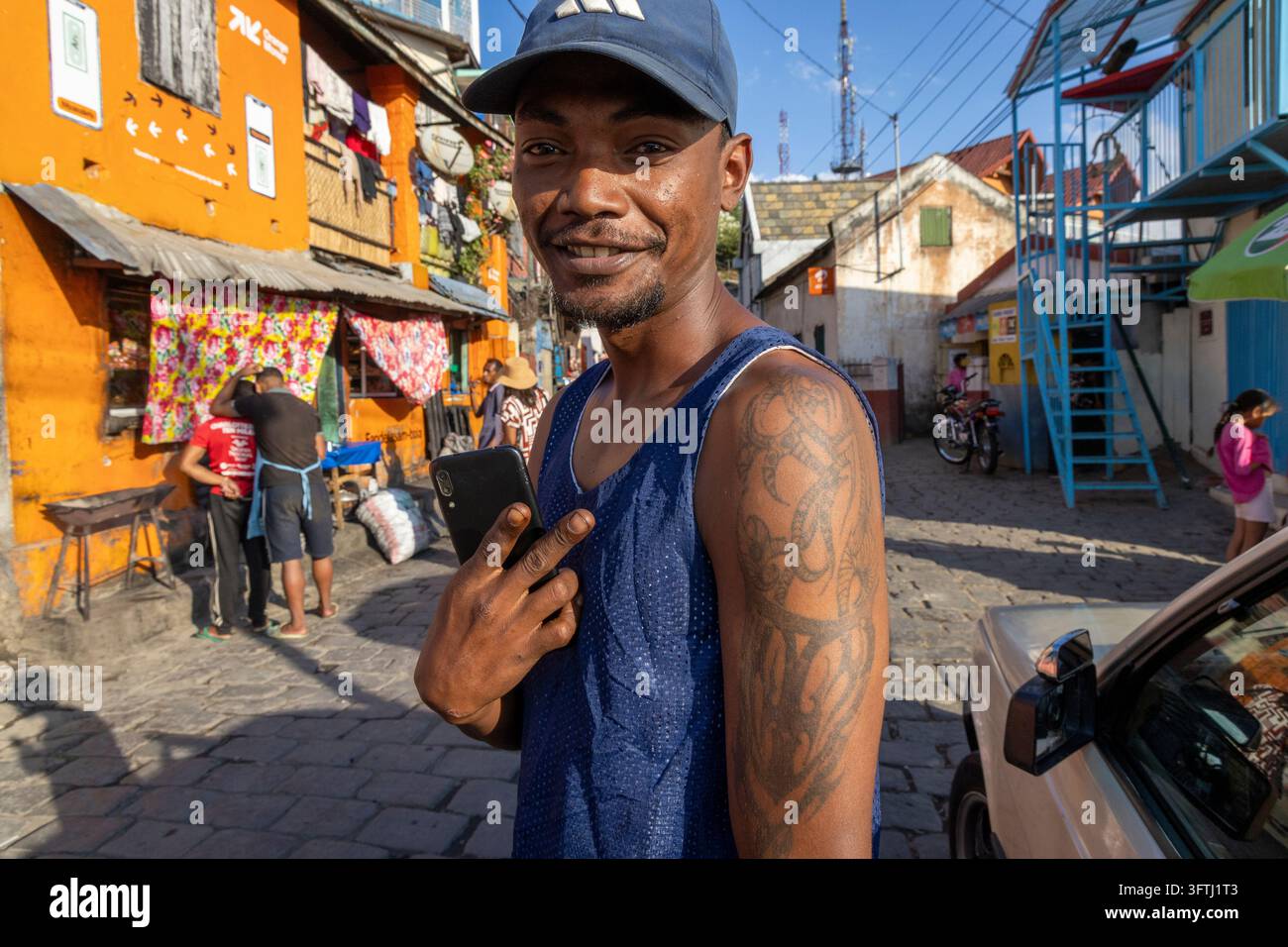 Giovane uomo sorridente con un braccio tatuato che tiene un telefono in una strada colorata, Analamanga, Antananarivo, Madagascar Foto Stock