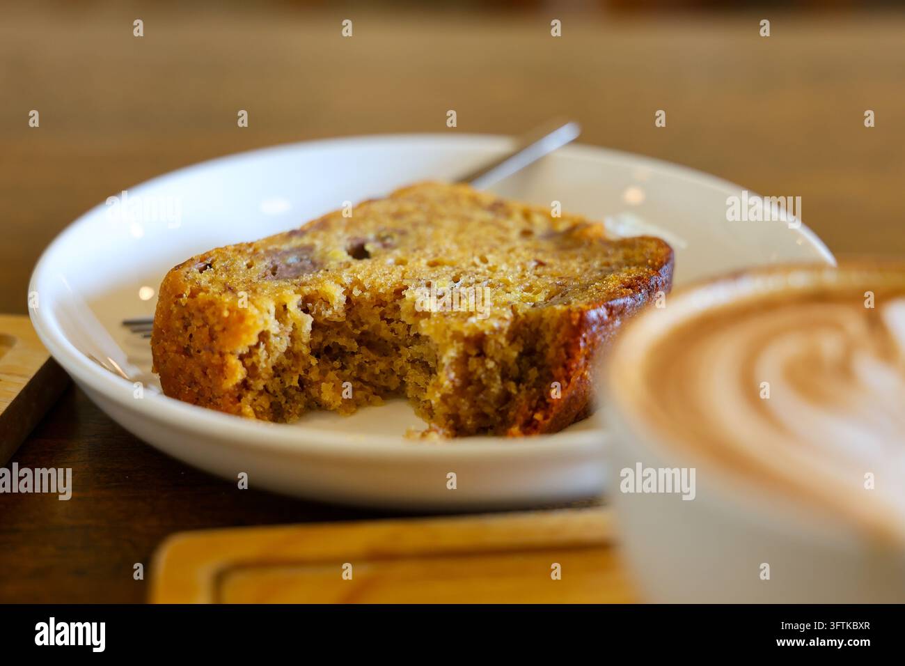 Una fetta di pane bagnato alle banane su un piatto bianco accanto a un latte in un ambiente da caffè, con crosta dorata, mollica morbida, tavolo di legno caldo e una colazione invitante a Foto Stock