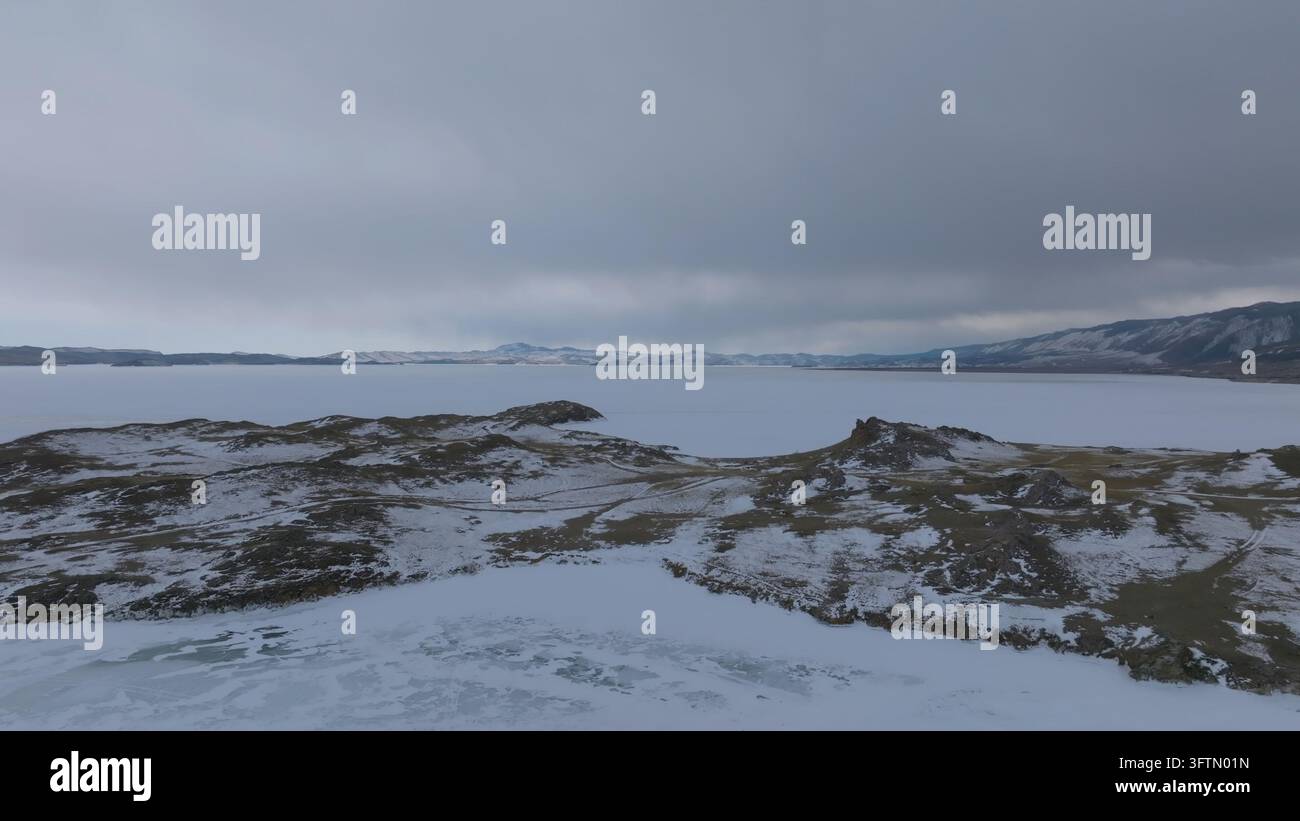 Una vista invernale elevata cattura il lago Bajkal accanto a Maloye More, dove creste innevate incorniciano la riva ghiacciata sotto un cielo smorzato nell'oblast di Irkutsk Foto Stock