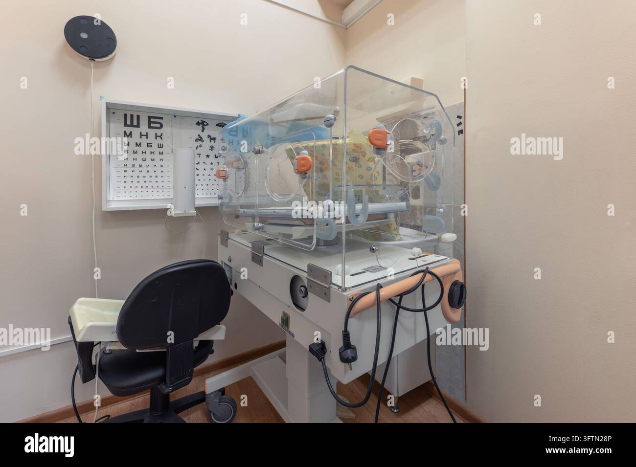 Un incubatore neonatale si trova accanto a una sedia e a tabelle di controllo della vista montate a parete in una stanza d'ospedale Foto Stock