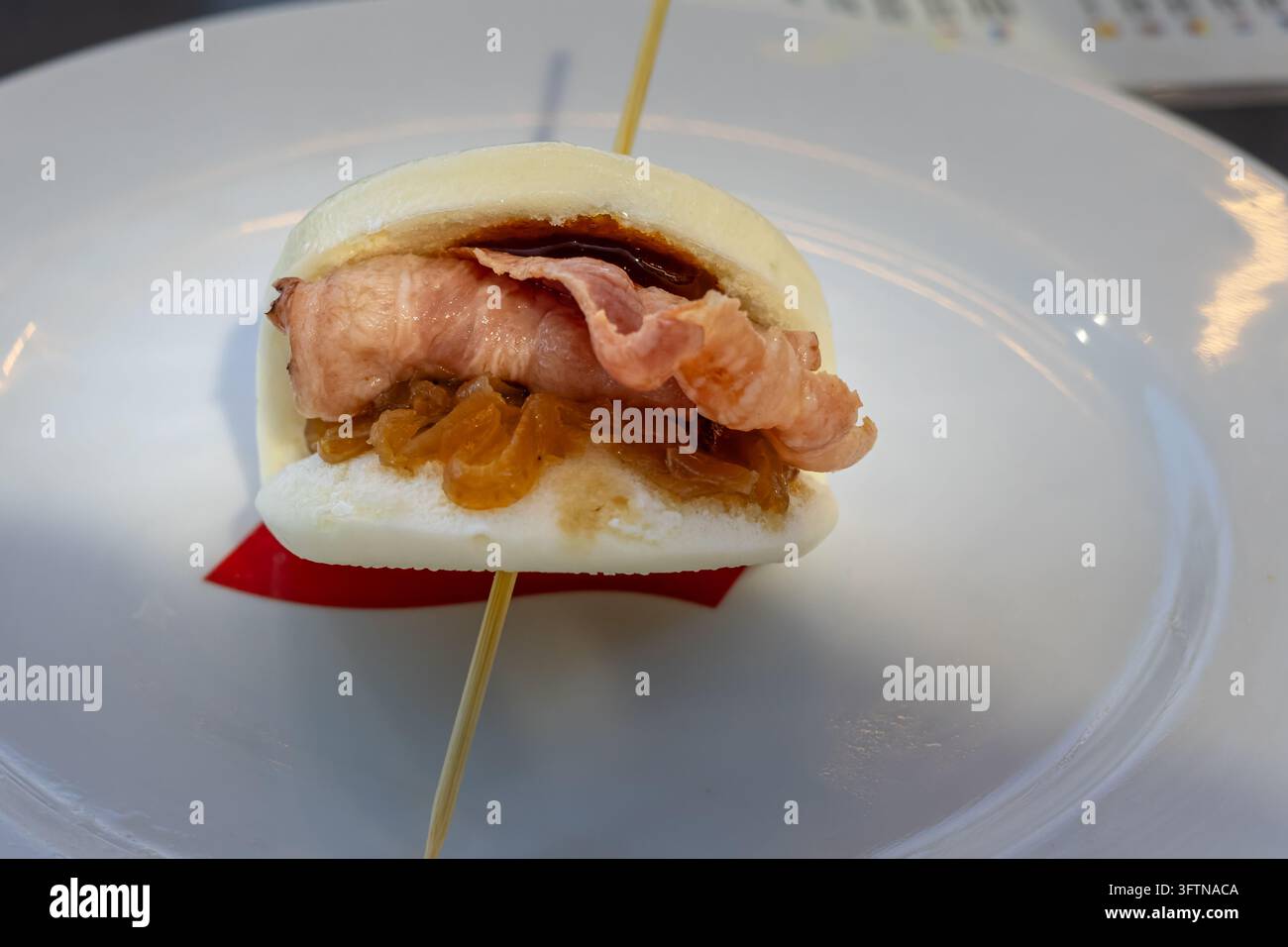 Prima vista di un bao bun al vapore ripieno di pancetta e chutney di cipolle, servito come antipasto gourmet su un piatto bianco Foto Stock
