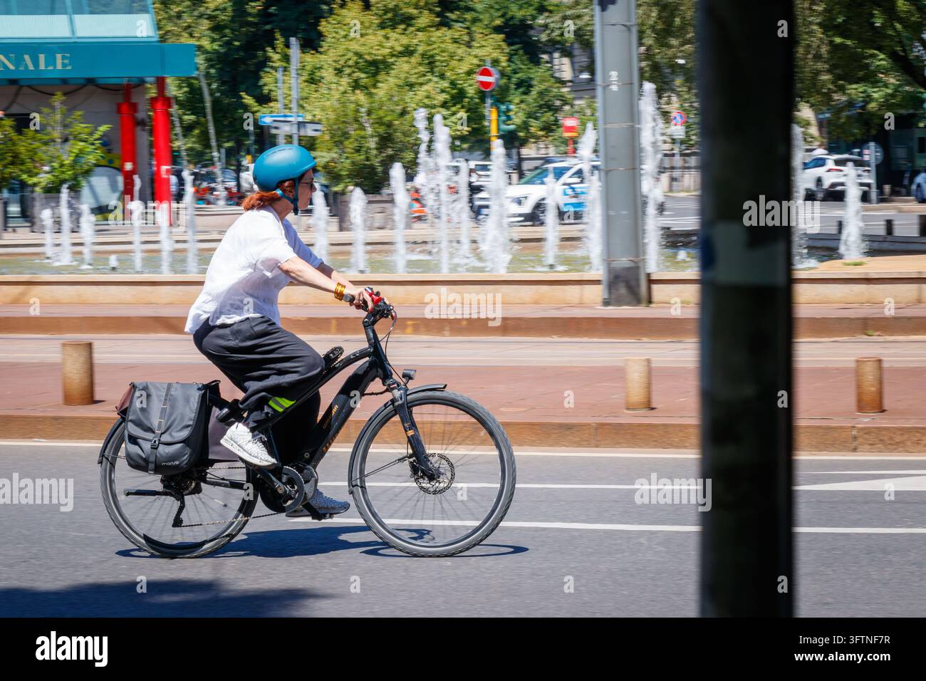 Ciclista in sella a una bicicletta elettrica nera con casco e borsa a pannello lungo una strada cittadina con sfondo di fontana Foto Stock