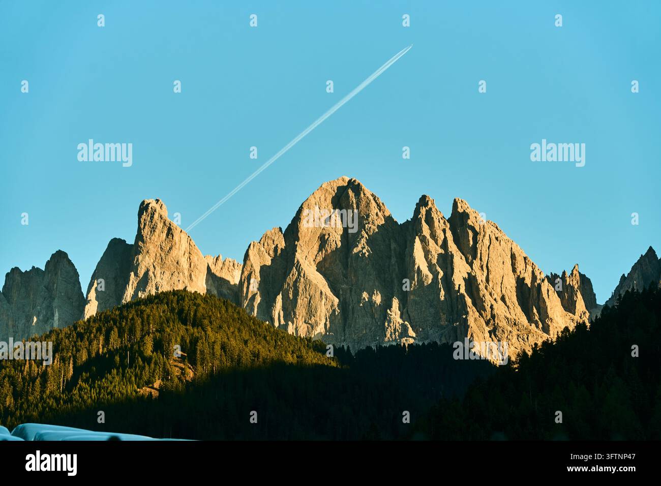 Picchi frastagliati delle Dolomiti bagnati da luce dorata sopra una foresta di conifere con cielo blu limpido e scia di condensa di un aereo che si estende sopra, alpino drammatico Foto Stock