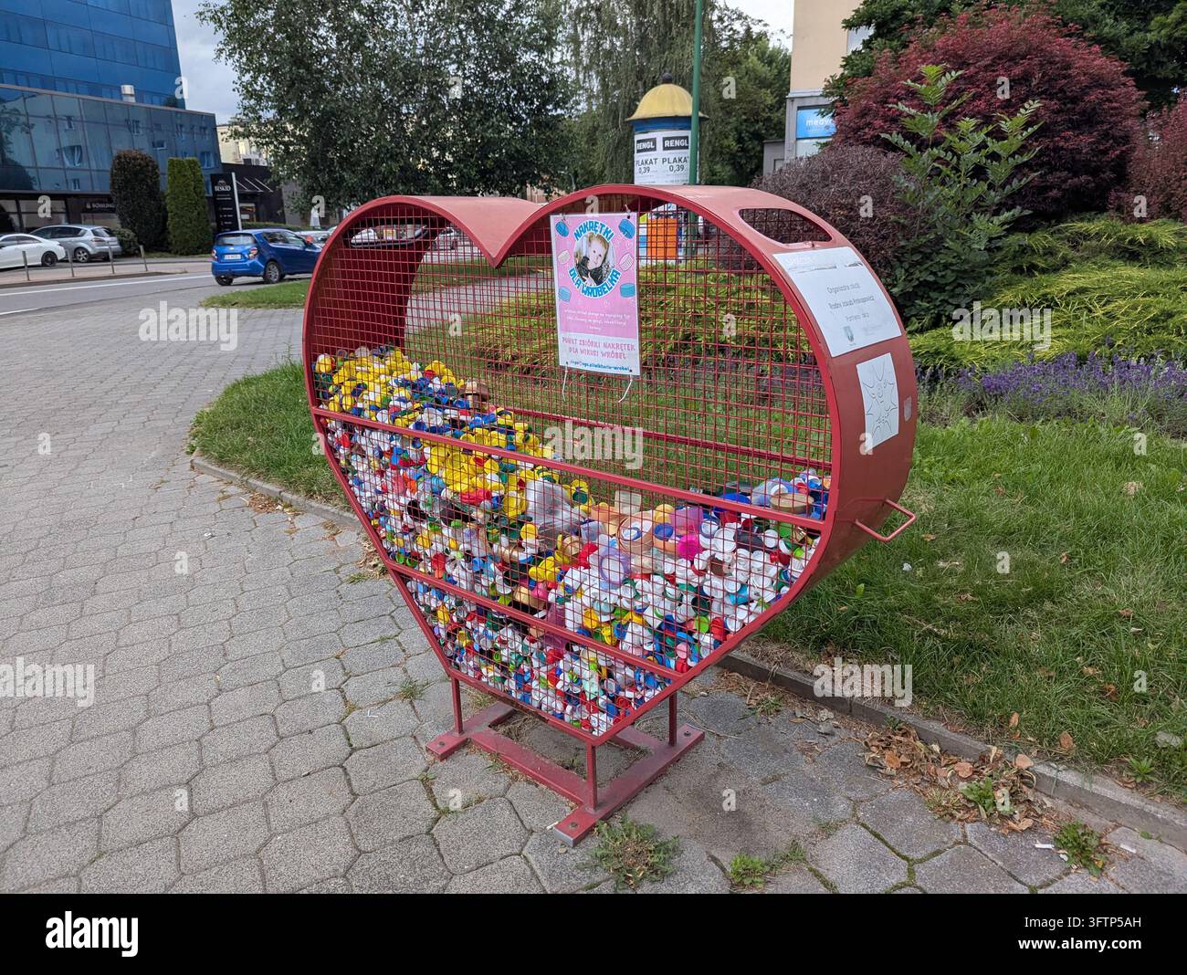 10 luglio 2026, Nowy Sacz, Polonia, Un bidone di riciclaggio a forma di cuore è pieno di tappi di plastica colorati, situato in un'area di parco urbano. Il bidone è fatto di Foto Stock