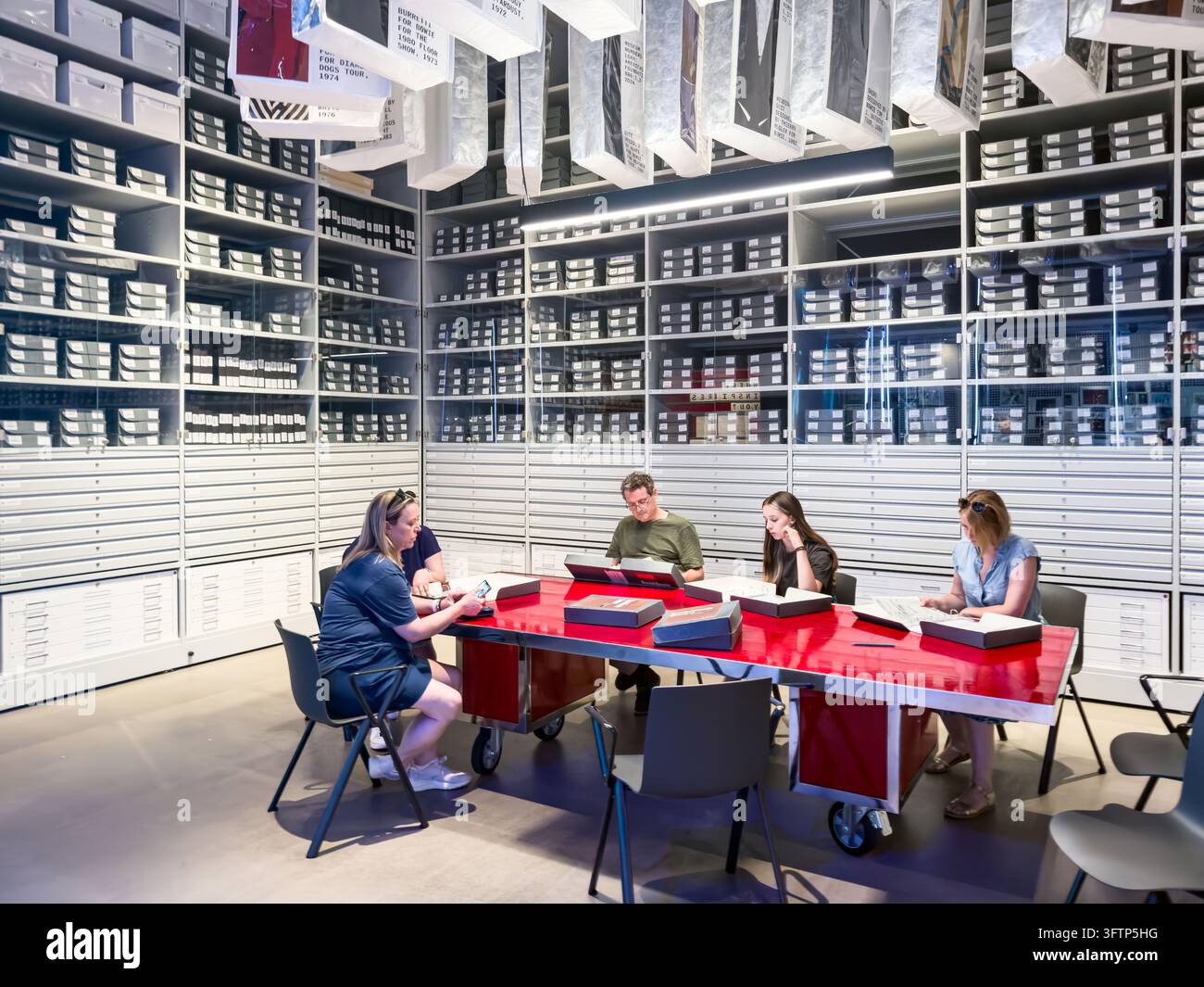 Visitatori seduti a un tavolo rosso che esaminano oggetti nella sala di studio dell'archivio con scaffali e scatole di archiviazione mostra David Bowie V&A East - Immagine stock catturata con smartphone