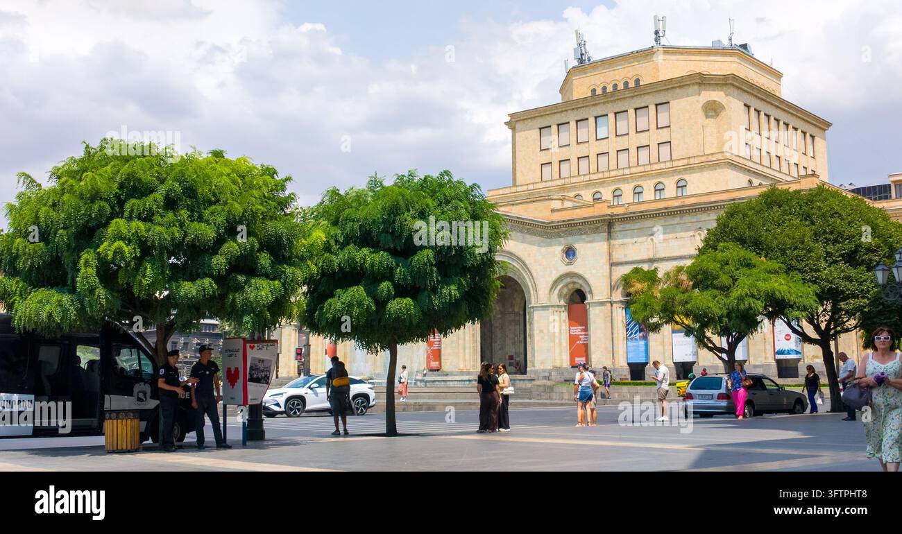 Grandi alberi verdi frondosi delimitano una piazza pubblica. Le persone camminano e si fermano vicino a un grande edificio storico. L'edificio ha archi, colonne e più wi Foto Stock