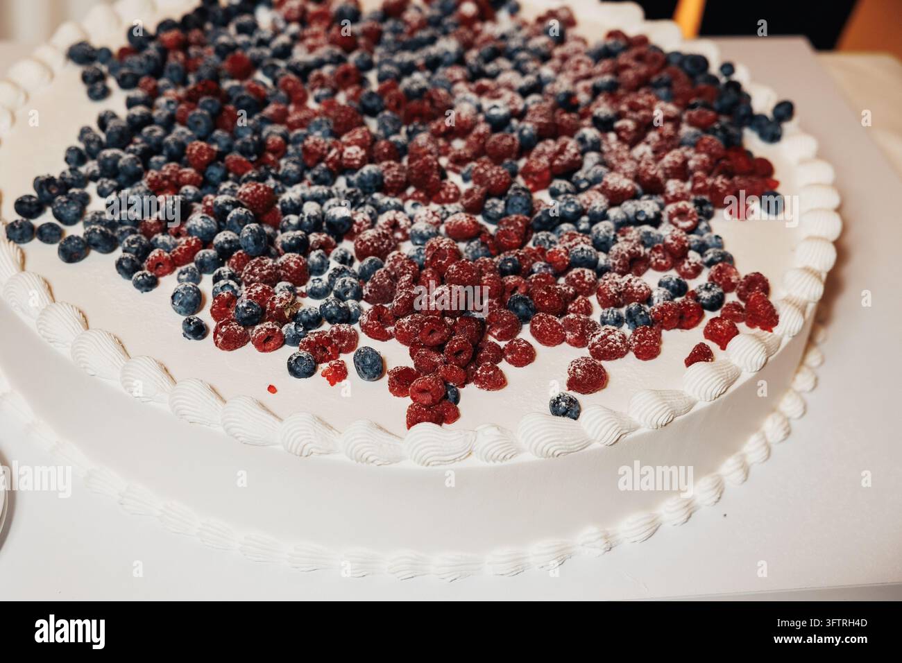 Torta rotonda bianca glassata ricoperta di lamponi e mirtilli spolverati di zucchero a velo, primo piano che mostra il bordo glassato decorato e la glassa liscia Foto Stock
